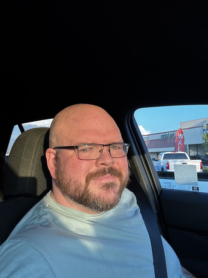 Sly Panorama selfie in the driver seat of a car wearing glasses and a seatbelt, daylight in a parking lot.