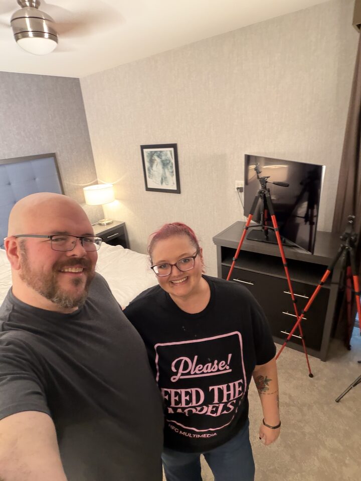 Sly Panorama hotel bedroom selfie with a smiling collaborator in a Please Feed The Models tee beside a lit tripod and TV.