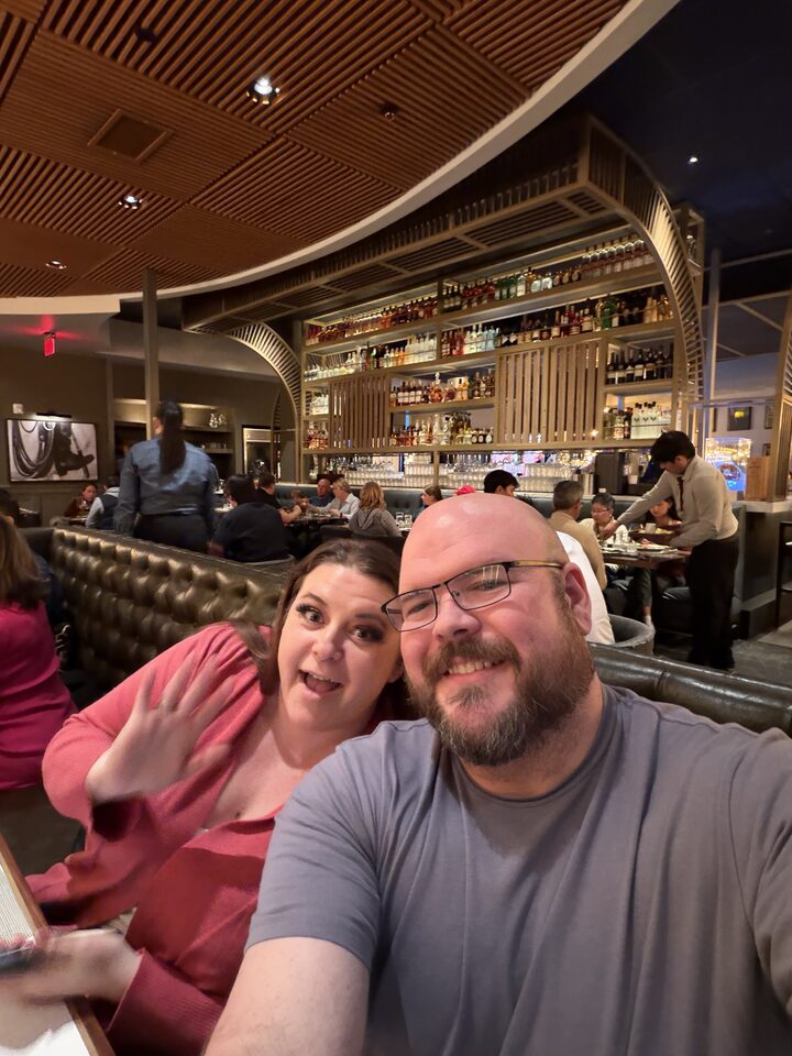 Sly Panorama selfie with a waving collaborator at a candlelit lounge bar with a fully stocked back bar.