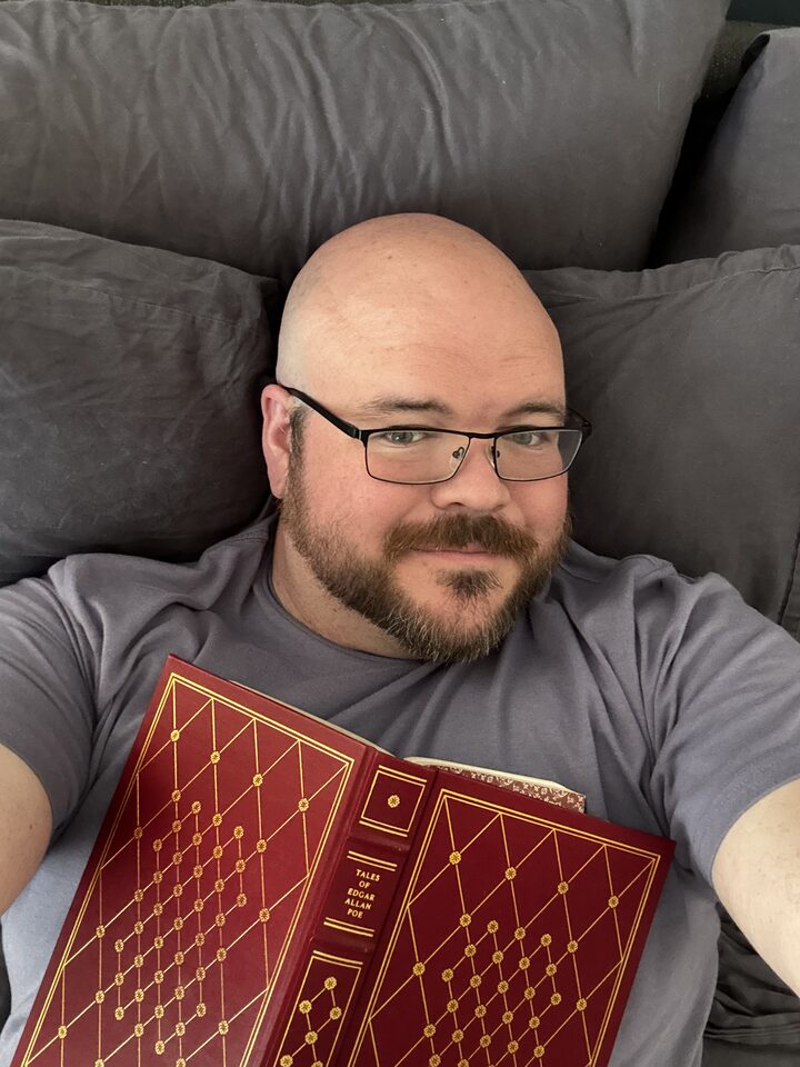 Sly Panorama reclining on a grey couch reading a leather-bound Tales of Edgar Allan Poe.