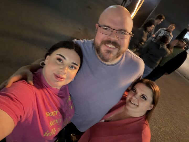 Sly Panorama in a periwinkle tee posing with two smiling collaborators at an adult industry expo.