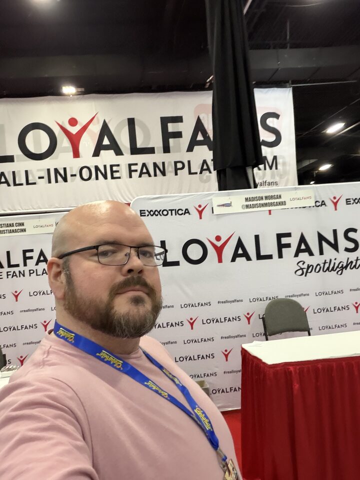 Sly Panorama selfie with a smiling collaborator beneath the JERKMATE balloon arch at Exxxotica.
