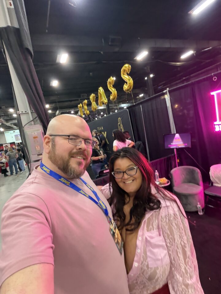 Sly Panorama solo selfie at the LoyalFans Spotlight booth at Exxxotica wearing a pink polo and event lanyard.