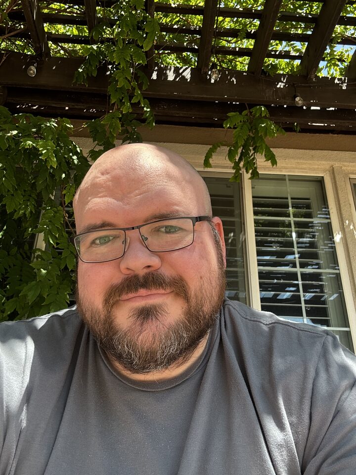 Sly Panorama daylight selfie in a grey tee beside a leafy garden trellis and window.