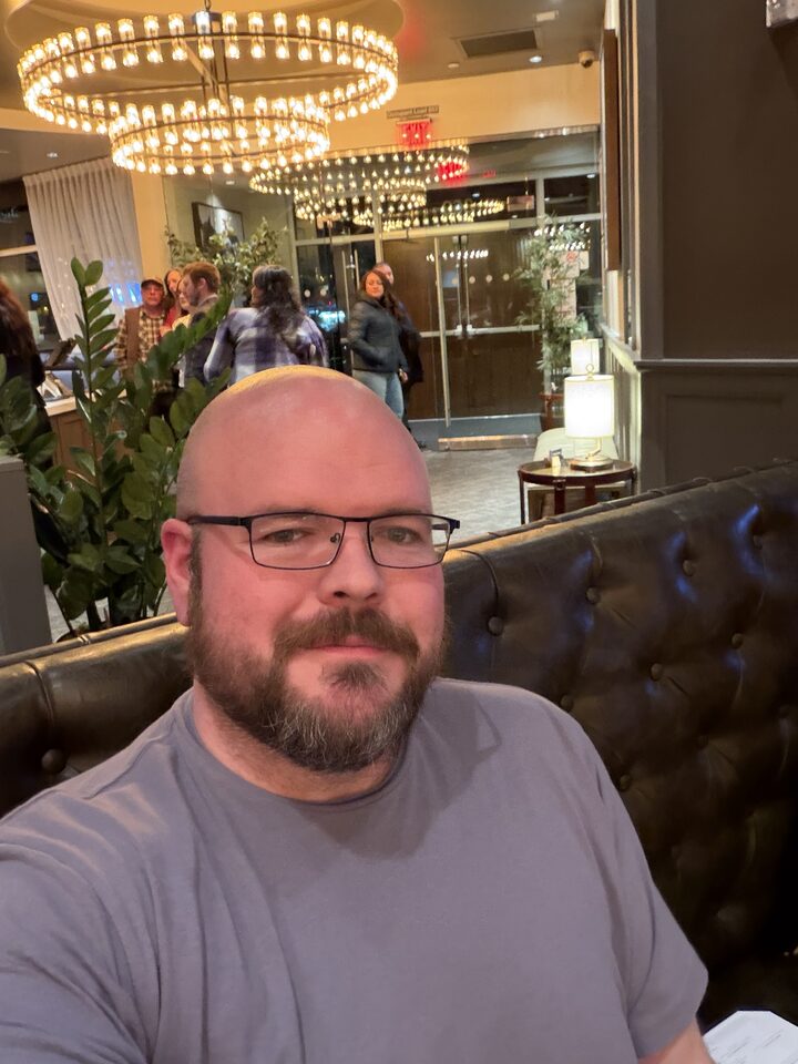 Sly Panorama selfie at a leather chesterfield booth under a beaded chandelier in an upscale pub.