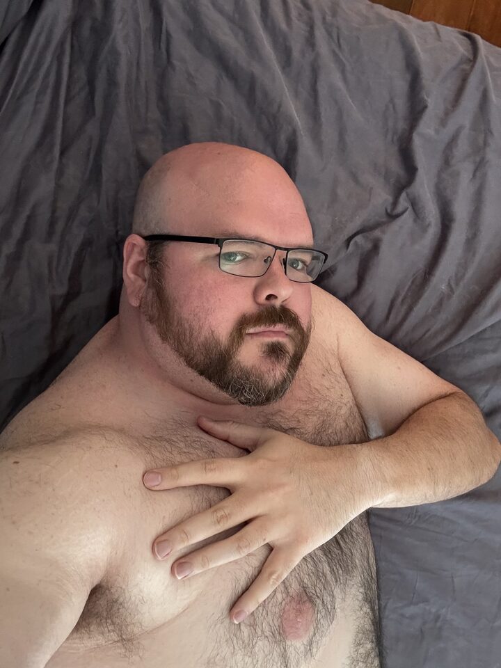 Sly Panorama shirtless on a bed with grey sheets, hand on chest, looking at the camera.