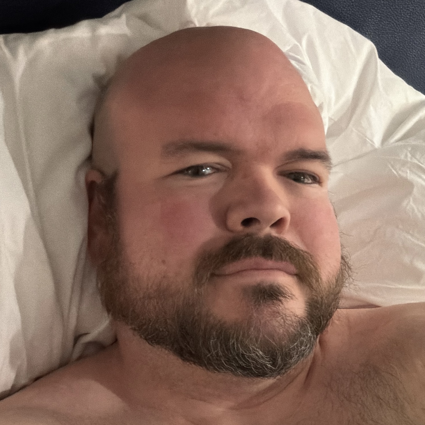Sly Panorama close-up shirtless selfie on white pillows in soft bedroom light.