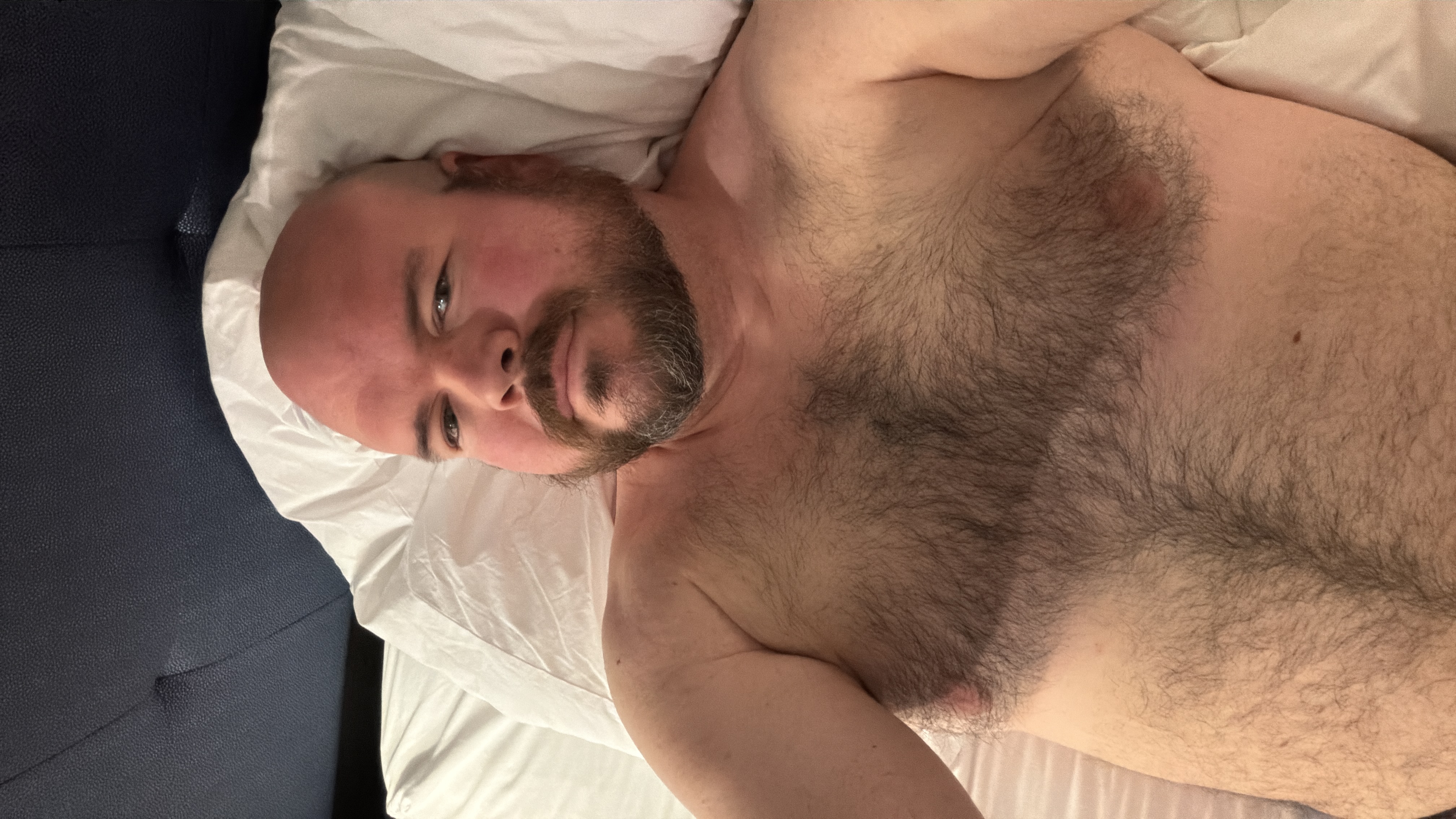 Sly Panorama shirtless on white pillows with a tufted dark headboard behind him.