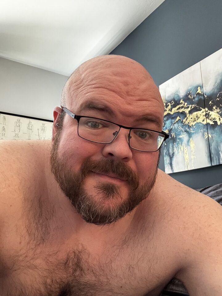 Sly Panorama shirtless selfie at home with blue wall and swirling blue, white, and gold artwork.