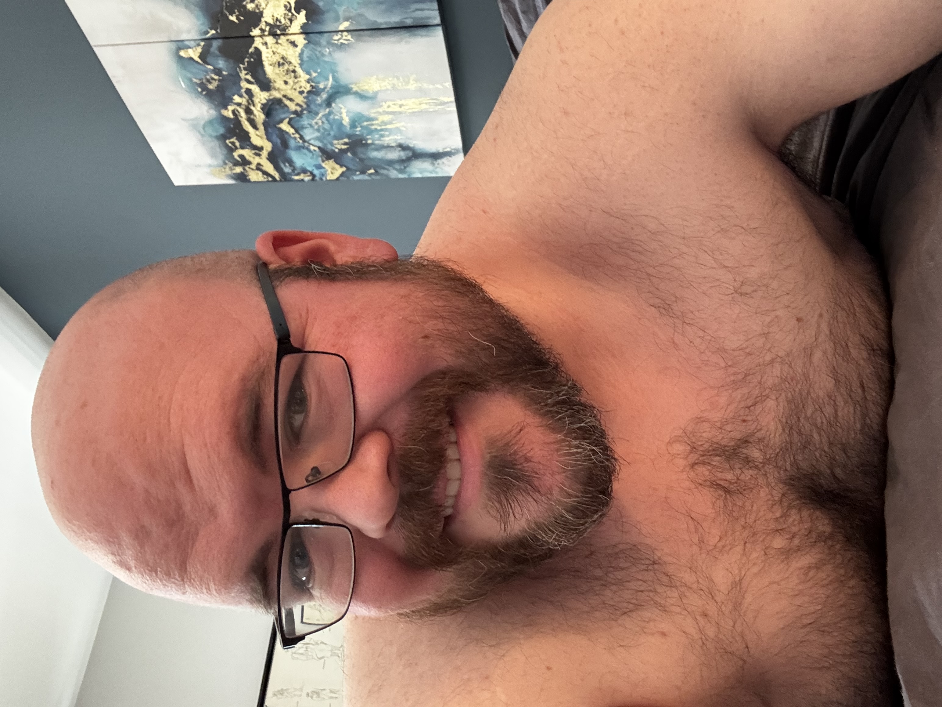 Sly Panorama shirtless, smiling, with blue and gold abstract art on the wall behind him.