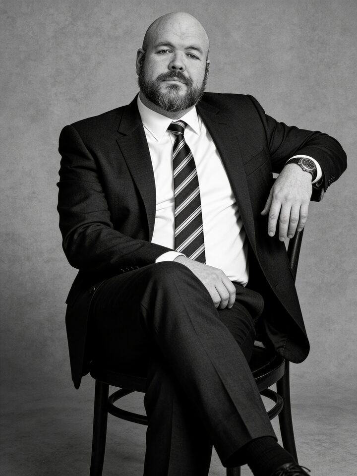 Sly Panorama black and white studio portrait with arm resting on the chair back, charcoal suit and striped tie.