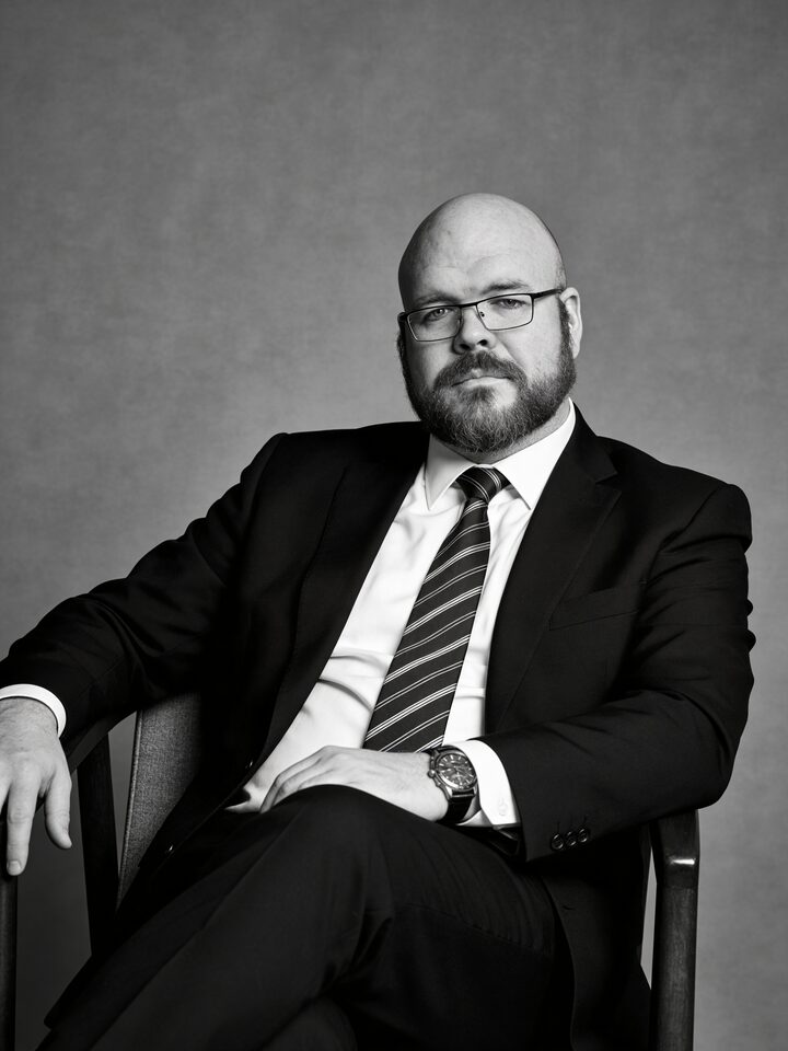 Sly Panorama black and white studio portrait seated with crossed leg in a charcoal suit, striped tie, and dress watch.