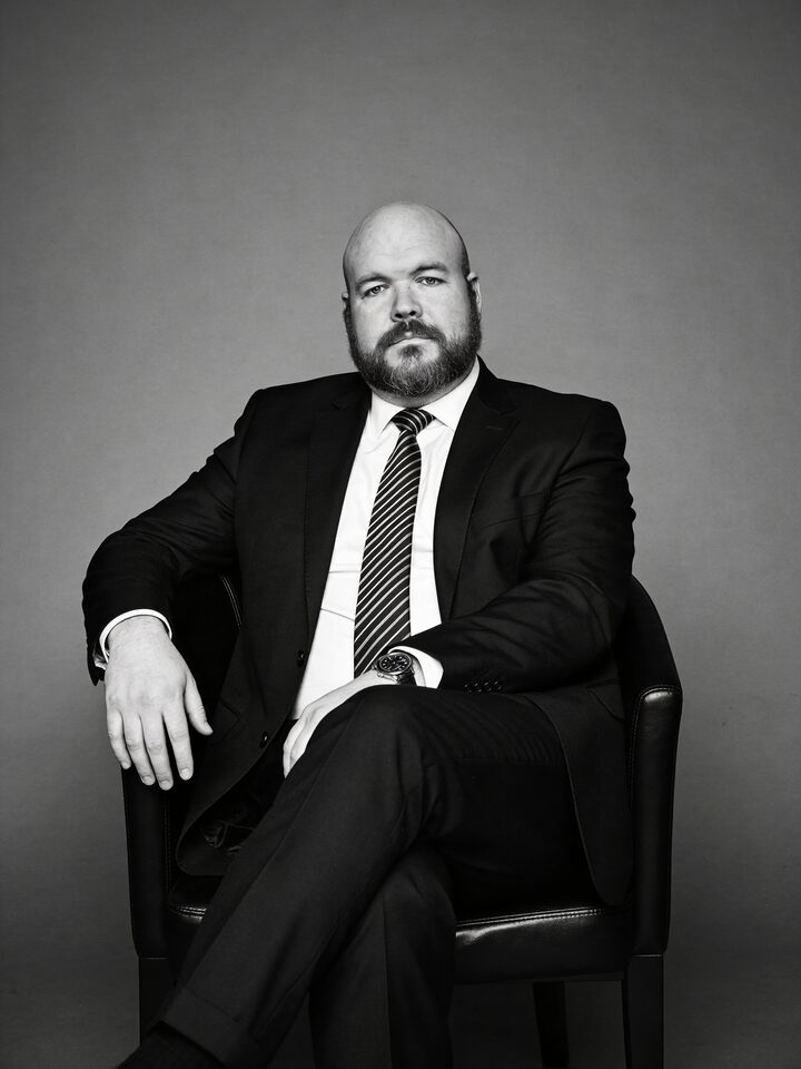 Sly Panorama black and white studio portrait relaxed in a chair wearing a charcoal suit and striped tie.