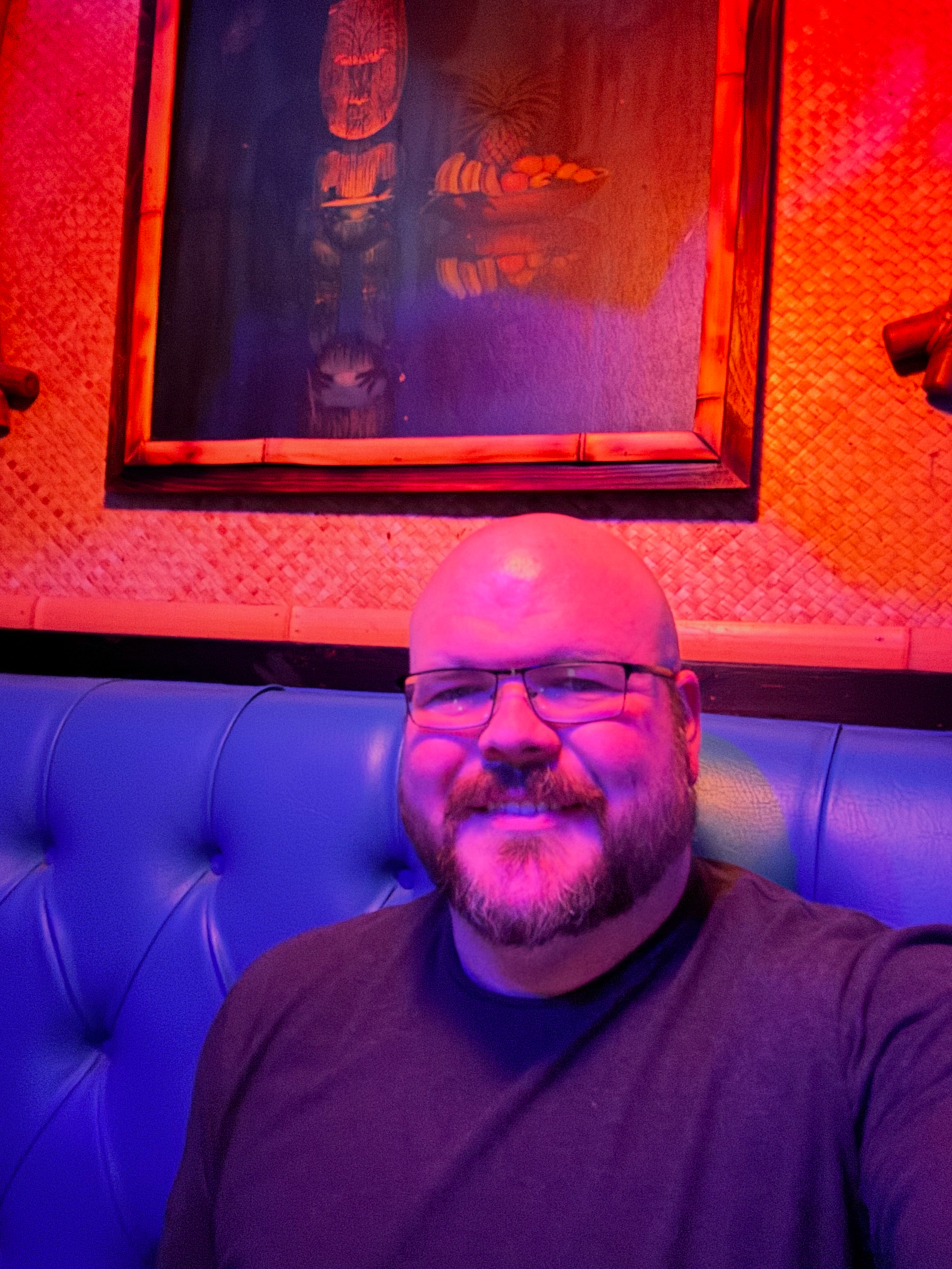 Sly Panorama smiling in a tiki-style bar seated in a bright blue booth with tropical wall decor.