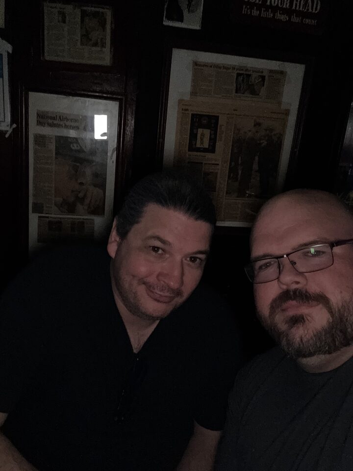 Sly Panorama selfie with a friend in a dim pub with framed memorabilia on the walls behind them.
